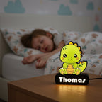 Veilleuse NextLab Studio personnalisée en forme de dinosaure vert sur une table de chevet près d'un enfant endormi.